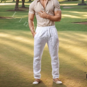 May include: A person wearing a short-sleeved tan button-down shirt and white drawstring pants. The person is standing in a park with green grass and trees. The person is wearing brown shoes. The image has a soft, natural lighting.