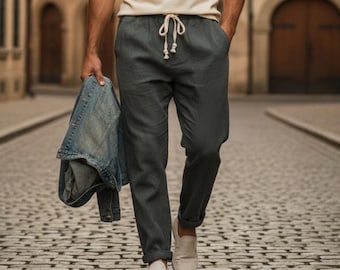 Pode incluir: Um homem vestindo uma camisa polo creme, calças cinza com cordão e sapatos brancos sem cadarços caminha por uma rua de paralelepípedos. Ele carrega uma jaqueta jeans. O fundo apresenta edifícios com telhados de telhas vermelhas.