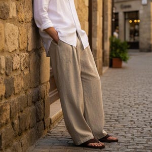 May include: A white linen shirt and beige linen trousers. The person is leaning against a stone wall. The outfit is paired with brown sandals. The background shows a cobblestone street and buildings.