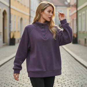 May include: A purple fleece pullover with a high collar and three dark buttons. The long-sleeved top has a relaxed fit and ribbed cuffs. The model is wearing black pants. The background is a cobblestone street with colorful buildings.