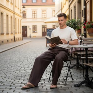 May include: A person seated outdoors, reading a book. They wear a cream-colored t-shirt, dark brown pants, and sandals. The scene is set on a cobblestone street with buildings in the background.