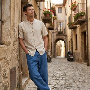 May include: A beige short-sleeved shirt with a mandarin collar and button closures, paired with blue denim jeans. The shirt has a relaxed fit. The person is leaning against a stone wall in a European street.