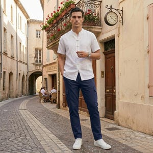 May include: A man wearing a white short-sleeve shirt with a mandarin collar and navy blue cargo pants. He is also wearing a silver watch and white sneakers. The background is a European street scene.