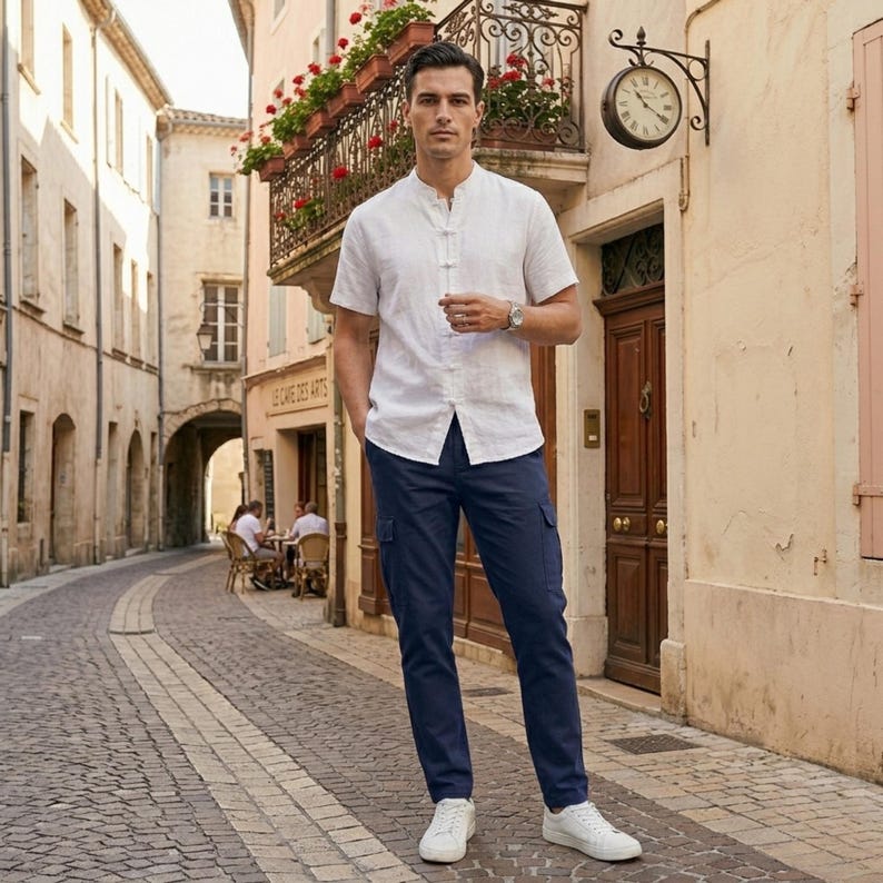 May include: A man wearing a white short-sleeve shirt with a mandarin collar and navy blue cargo pants. He is also wearing a silver watch and white sneakers. The background is a European street scene.