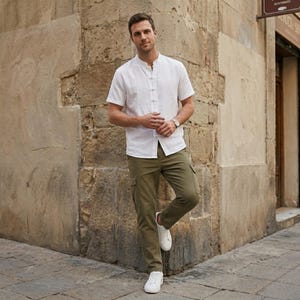 May include: A person wearing a white short-sleeve shirt with a mandarin collar and olive green cargo pants. The person is also wearing a silver watch and white sneakers. The person is leaning against a stone wall.