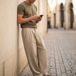 May include: A person wearing an olive green short-sleeve t-shirt and beige pleated trousers. The person is leaning against a light-colored wall and holding a black smartphone. The background features a cobblestone street and arched doorways.