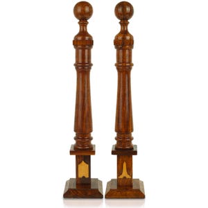 May include: Two tall, ornate wooden candle holders. Each has a round top, a long, turned column, and a square base. The wood is a rich brown color with lighter wood accents. The candle holders are symmetrical and stand upright.