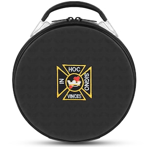 May include: A black, circular carrying case with a top handle and zip. The case has a gold and yellow emblem with the text "HOC SIGNO VINCES" and "IN". The handle is black with silver detailing. The case has a textured pattern.