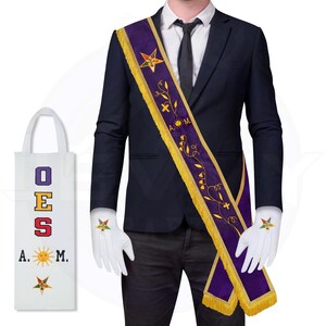 May include: A purple sash with gold fringe and embroidered floral details is draped over a dark suit. White gloves with star emblems and a white tote bag with colorful text and symbols are also visible. The sash has the letters "A.M."