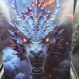 May include: A rectangular print featuring a detailed dragon head with iridescent blue and purple scales, glowing red eyes, and sharp horns. The dragon is depicted with its mouth slightly open, surrounded by dark clouds and floating orange embers.