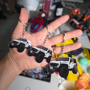 Könnte beinhalten: Drei weiße Jeep Rubicon SUV-Schlüsselanhänger mit schwarzen Reifen, einem schwarzen Kühlergrill und einem silbernen Metallring mit Kette. Jeder Schlüsselanhänger zeigt eine detaillierte Grafik eines weißen Geländewagens mit dem Rubicon-Schriftzug.