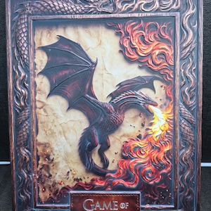 Puede incluir: Una placa de pared rectangular con un relieve de un dragón escupiendo fuego. El marco está decorado con un cuerpo de dragón y llamas. El centro muestra un dragón en vuelo sobre un fondo de pergamino. El texto GAME OF THRONES está abajo.