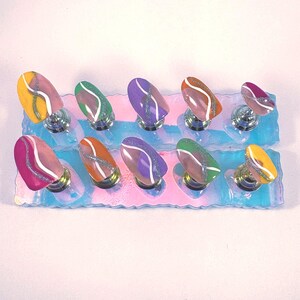 May include: A set of ten oval-shaped false nails in various colors, including yellow, green, purple, and pink. Each nail features a white and silver glitter design. The nails are displayed on a clear, iridescent stand.