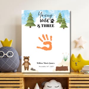 May include: A framed print with the text "Young, Wild & Three" and an orange handprint. The print features a watercolor sky and pine trees. Below the handprint is the name "Willow Marie James" and the date "November 16th, 2025".