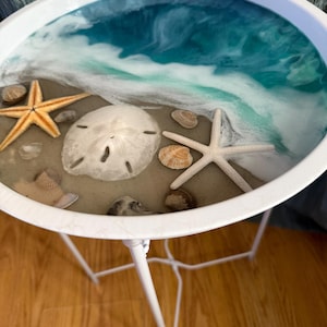 Pode incluir: Uma mesa lateral redonda com estrutura de metal branco e tampo com design em resina representando uma cena de praia. A superfície exibe areia, um grande dólar-da-areia branco, duas estrelas-do-mar e várias conchas pequenas.