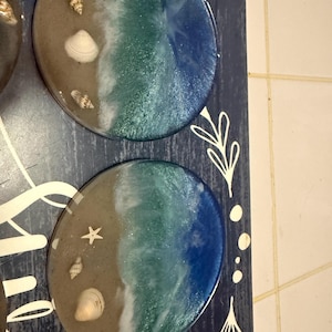Puede incluir: Posavasos de resina con temática costera. Cada posavasos presenta un diseño de playa arenosa con conchas marinas y un efecto de ola en tonos azules. Los posavasos están sobre una superficie de madera azul oscuro.
