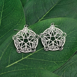 May include: A pair of silver-colored earrings with a pentagram design. The earrings feature intricate cut-out patterns of stars, geometric shapes, and floral elements. The earrings are displayed on a background of green leaves.