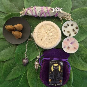 May include: A collection featuring a sage bundle with purple twine and a silver charm, three incense cones on a black plate, a round textured disc, two tea light candles with crystals, a purple velvet pouch, and a black cat illustration tin.