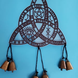 May include: A brown wooden wall hanging in a Celtic knot design with the words "Speak with Love", "Honor your Word", and "Be Balanced". It has five bronze bells hanging from black strings. The center has a pentagram.