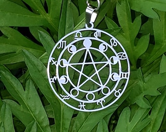 Puede incluir: Un collar con colgante plateado con un diseño circular con símbolos astrológicos y un pentagrama central. El colgante cuelga de un cordón negro. El diseño incluye fases lunares. El fondo son hojas verdes.