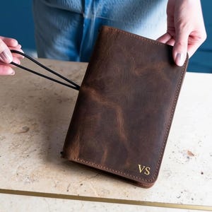 May include: A dark brown leather notebook cover with a distressed finish and visible grain texture. The cover features the gold embossed initials VS in the bottom right corner and includes a thin black elastic cord attached to the side for closure.