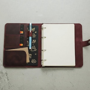 Puede incluir: Carpeta de cuero burdeos abierta con múltiples bolsillos, ranuras para tarjetas y un portalápices. La carpeta tiene un bloc de notas en blanco, un inserto negro con ilustraciones doradas y un cierre a presión. Hay dos bolígrafos en el portalápices.