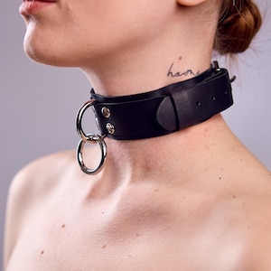 May include: A black leather collar with a silver metal ring detail. The collar is worn around the neck and has a buckle closure. The word "home" is tattooed on the neck.