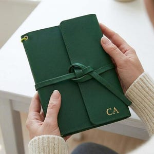 Può includere: Un diario in pelle verde scuro con chiusura a cinturino avvolgente annodato sul davanti. La copertina è personalizzata con le iniziali CA impresse in oro nell'angolo in basso a destra e include un cordino elastico color verde lime brillante.