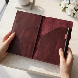 May include: A deep burgundy leather notebook cover featuring an open design with two interior pockets for document storage and a dedicated leather loop on the right side holding a slim black pen. The interior texture shows a natural matte finish.