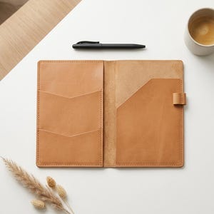 Pode incluir: Uma capa de caderno em couro castanho claro com acabamento exterior liso, dois bolsos interiores com bordas em forma de chevron no lado esquerdo, um bolso diagonal no lado direito e uma pequena presilha de couro para caneta na borda direita.