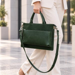 May include: A dark forest green rectangular briefcase featuring a smooth matte finish, two top handles, and a detachable shoulder strap. The bag includes a prominent front exterior pocket with a horizontal zip closure and silver-toned metal hardware.