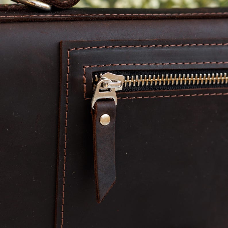 May include: Close-up of a dark brown leather bag with a gold zipper. The bag has a rectangular pocket with a zipper closure. The leather is stitched with brown thread. The zipper pull is a small piece of leather.