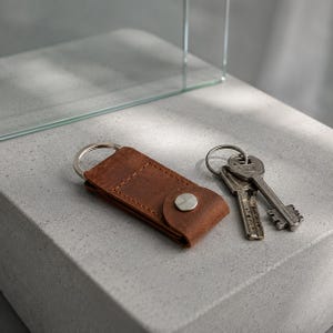 May include: A brown leather key organiser featuring a silver-toned metal snap button closure and a circular metal split ring. Two silver-coloured metal keys are attached to the ring, resting alongside the folded leather key holder accessory.