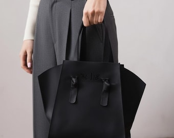 May include: A black tote bag with a structured design. The bag features two handles and decorative knots. The bag has a minimalist style and is held by a person wearing a watch and a white sweater.