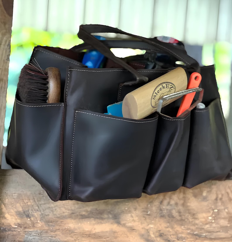 May include: A dark brown tote bag with multiple exterior pockets, filled with grooming tools. The bag has a handle and is made of a smooth material. Visible tools include a brush, a wooden cylinder with the text "SleekEZ", and an orange tool.