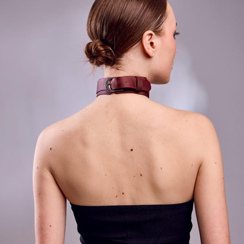 May include: A burgundy leather choker with a silver-colored buckle. The choker is worn around the neck. The background is a solid gray color. The person is wearing a black strapless top.