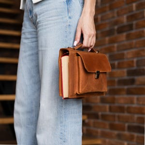 K&ouml;nnte beinhalten: Eine braune Ledertasche mit Klappenverschluss und oberem Griff. Ein dickes Buch mit roten Kanten wird in der Tasche gehalten. Die Tasche wird von einer Person getragen, die eine blaue Jeans tr&auml;gt. Der Hintergrund zeigt eine Backsteinmauer und Holztreppen.