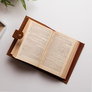 K&ouml;nnte beinhalten: Ein offenes Buch mit gealterten Seiten, eingebettet in einen braunen Lederumschlag. Der Umschlag hat einen Knopfverschluss. Das Buch liegt auf einem wei&szlig;en Hintergrund, mit etwas Gr&uuml;n in der oberen linken Ecke.