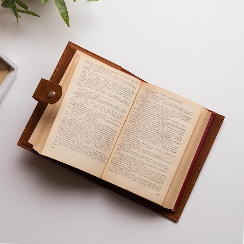 K&ouml;nnte beinhalten: Ein offenes Buch mit gealterten Seiten, eingebettet in einen braunen Lederumschlag. Der Umschlag hat einen Knopfverschluss. Das Buch liegt auf einem wei&szlig;en Hintergrund, mit etwas Gr&uuml;n in der oberen linken Ecke.