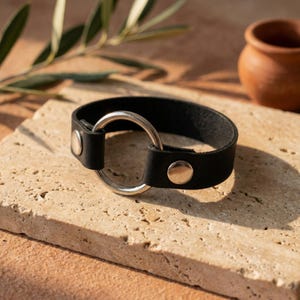 May include: A black leather bracelet featuring a circular silver-toned metal ring at the center. The leather band is secured on both sides of the ring with smooth, polished silver-toned metal rivets, creating a clean and functional design.