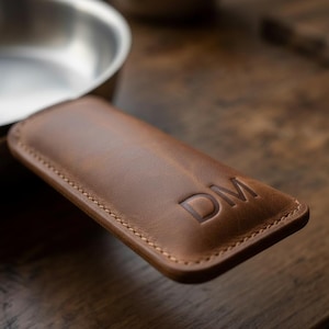 May include: A brown leather handle cover for a skillet, featuring visible stitching along the edges and the embossed letters DM on the surface. The cover is designed to fit over the metal handle of a cooking pan, providing a textured grip.