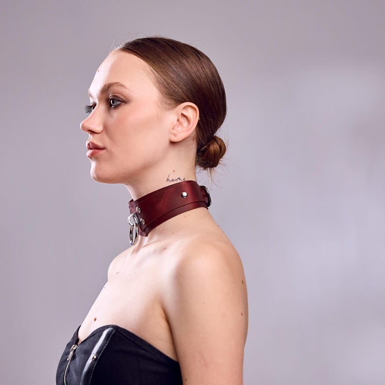 May include: A burgundy leather choker with silver-tone hardware. The choker features a wide band with a buckle closure and a large metal ring. The design is simple and elegant, suitable for various styles.