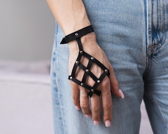 May include: Black leather hand harness with a grid design, featuring silver-tone rivets. The harness includes a wrist strap and a finger loop, creating a unique accessory. The accessory is worn on the hand.