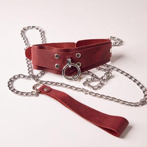 May include: A burgundy leather collar with a silver metal ring and rivets, connected to a silver chain leash. The collar is wide and has a layered design. The leash has a matching burgundy leather handle.