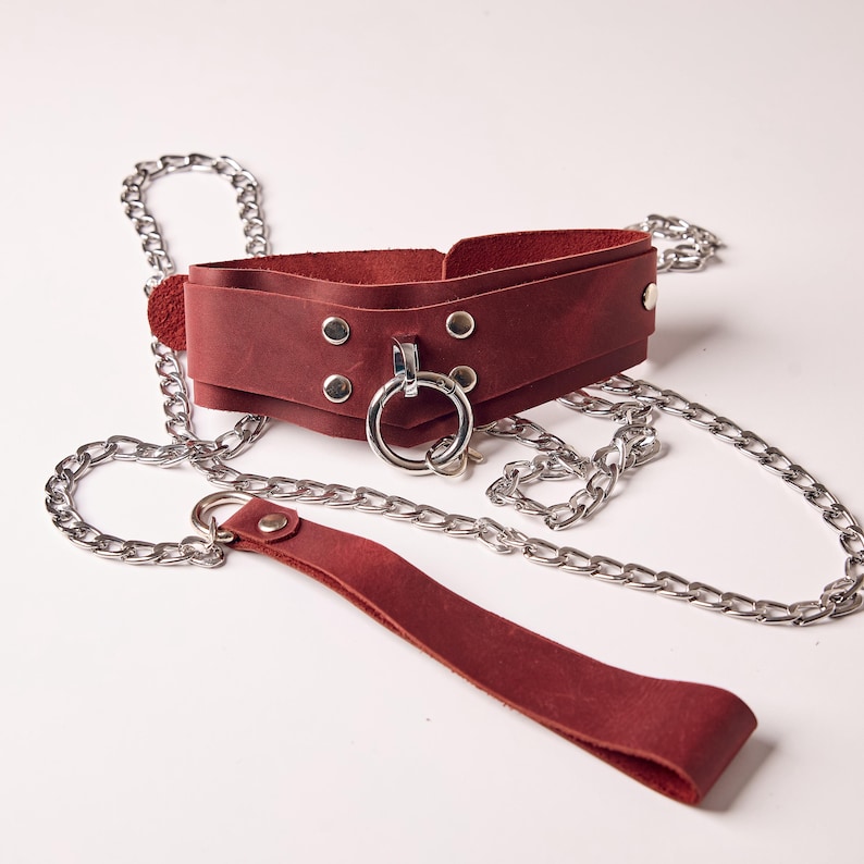 May include: A burgundy leather collar with a silver metal ring and rivets, connected to a silver chain leash. The collar is wide and has a layered design. The leash has a matching burgundy leather handle.