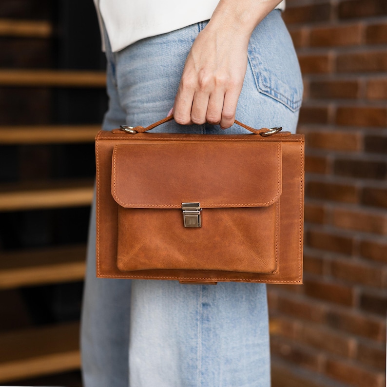 K&ouml;nnte beinhalten: Eine braune Lederaktentasche mit einem oberen Griff und einer Frontklappe, die mit einem Metallverschluss gesichert ist. Die Tasche hat eine rechteckige Form und eine Tasche auf der Vorderseite. Das Leder hat ein strukturiertes Aussehen.