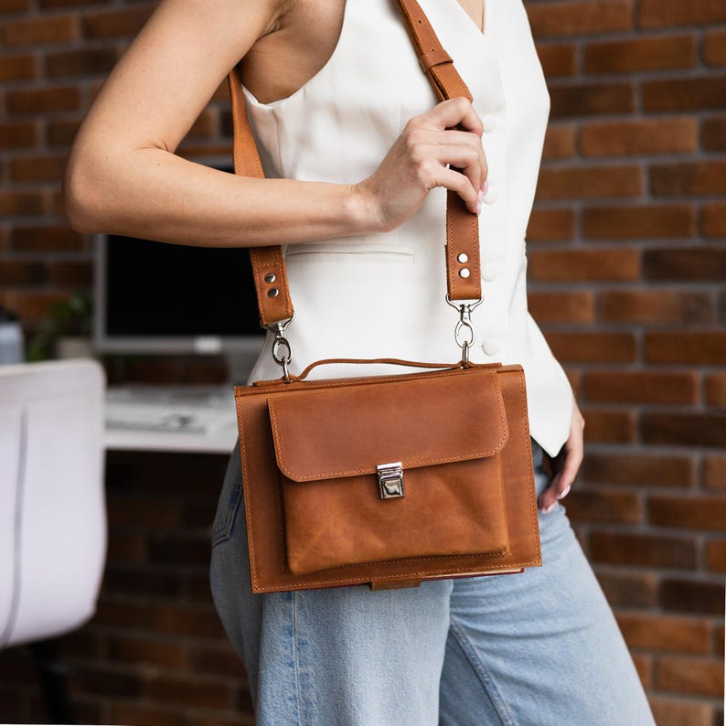 K&ouml;nnte beinhalten: Eine braune Leder-Umh&auml;ngetasche mit Klappenverschluss und silberfarbenem Verschluss. Die Tasche hat einen Schultergurt und einen Griff. Die Tasche ist aus hellbraunem Leder gefertigt.