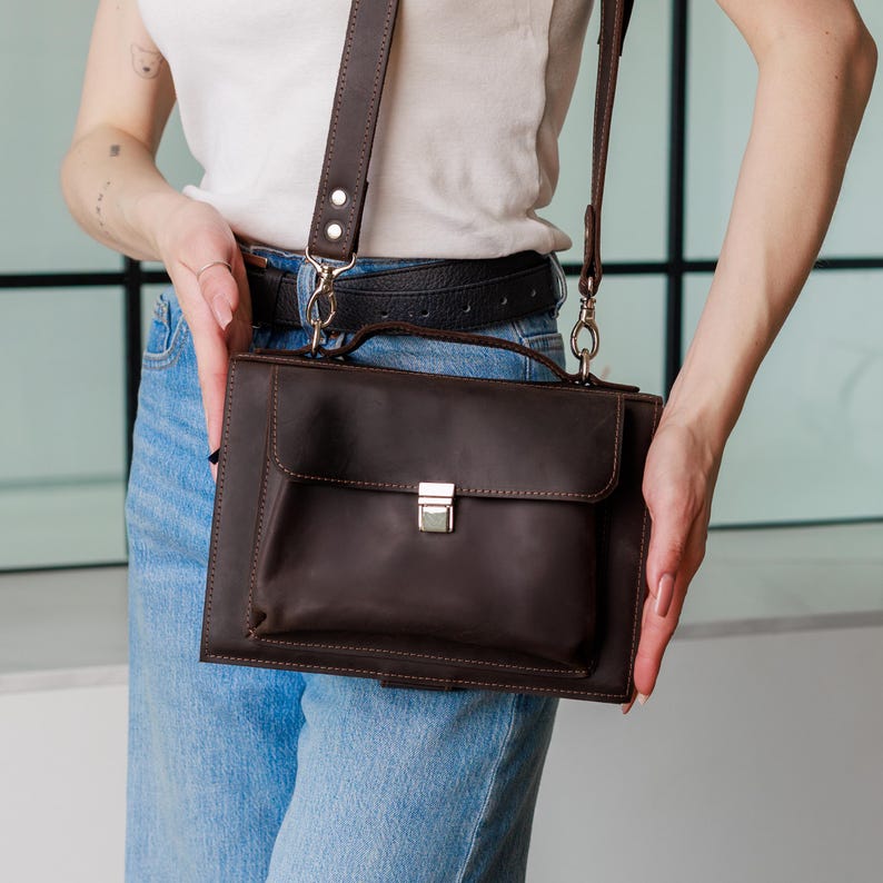 May include: A dark brown leather crossbody bag with a flap pocket and a silver-colored clasp. The bag has a matching brown leather strap and a handle. The bag is held by a person wearing a white shirt and blue jeans.