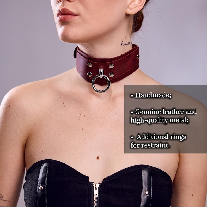 May include: A burgundy leather choker with a large silver ring and metal accents. The choker is handmade with genuine leather and high-quality metal, featuring additional rings for restraint.