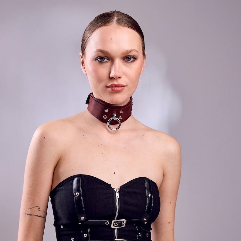 May include: A burgundy leather choker with a silver metal ring and studs. The choker is worn around the neck and has a buckle closure. The background is a solid gray.